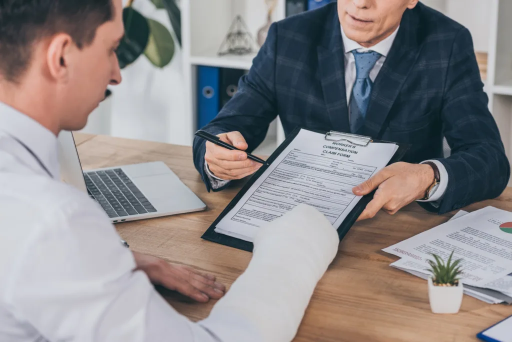 businessman in blue jacket giving form for compensation claim to worker with broken arm at table in